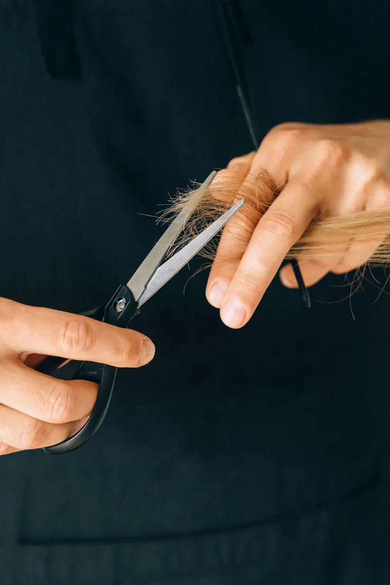 Nahaufnahme: zwei Hände führen eine Schere und eine helle Haarsträhne vor einer dunkelgrünen Schürze